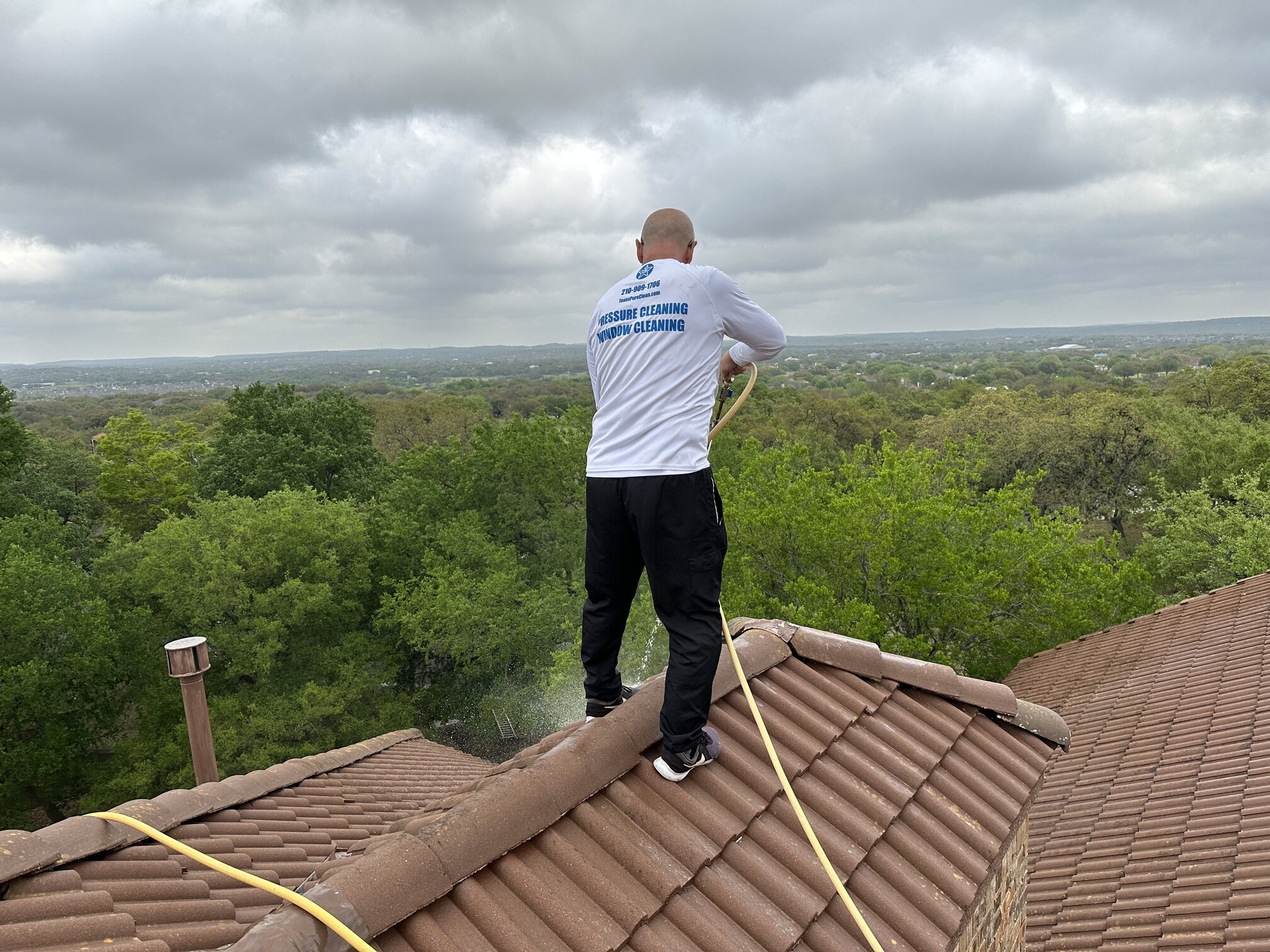 Owner Bill Eary standing on a tile roof in Fair Oaks Ranch, running the soft wash line — Hill Country tree line and clouds in the background