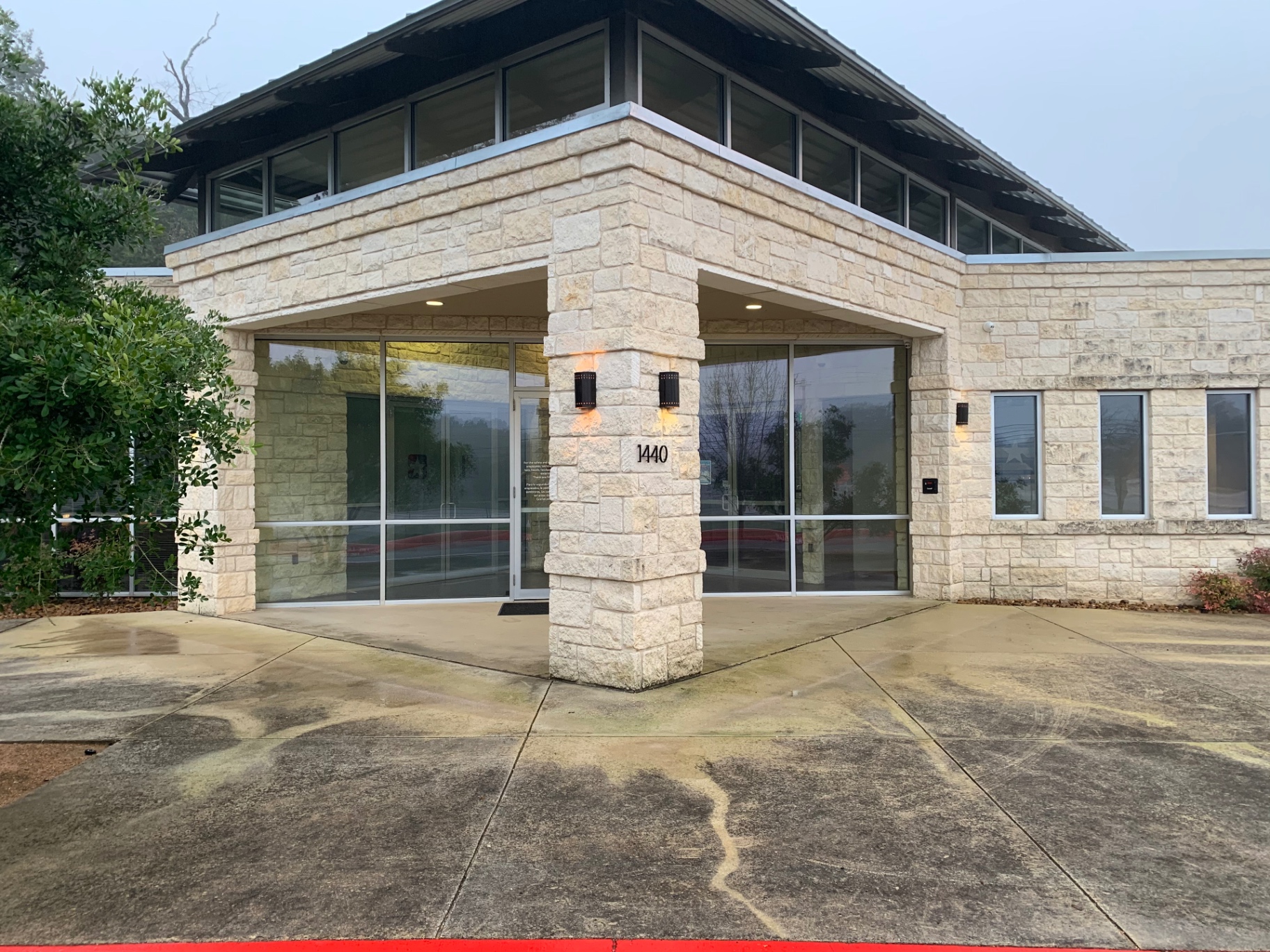 Front entry of the United Texas Credit Union Boerne branch before cleaning, showing green and orange organic staining across the concrete pad and stone columns
